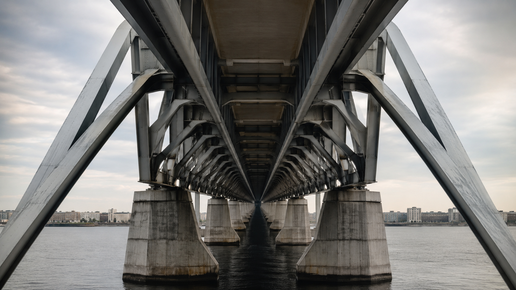 The underside of a long river bridge, its steel bracing and concrete piers receding symmetrically towards a distant vanishing point