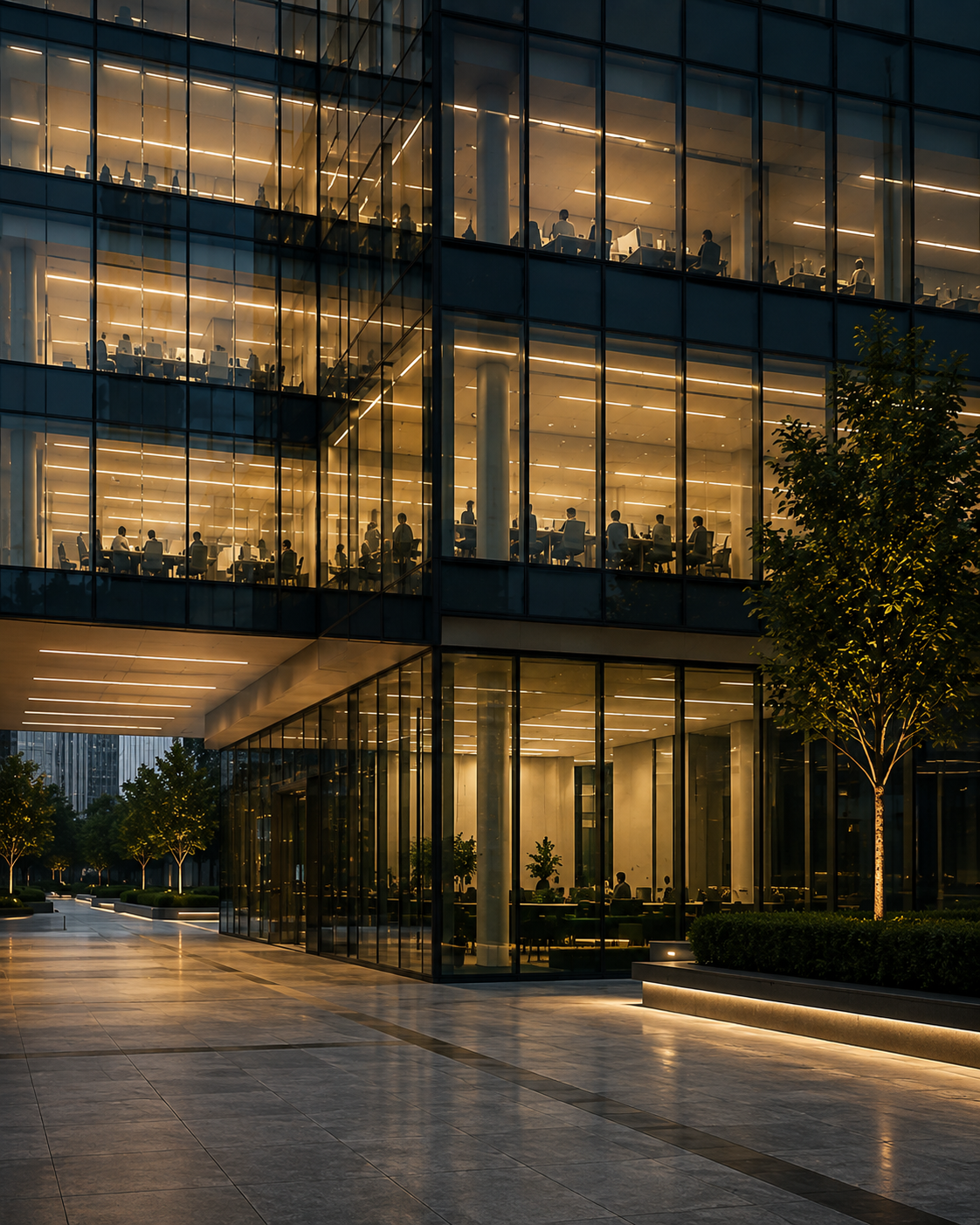 A lit office building at dusk, its occupied floors visible above a colonnade and paved forecourt